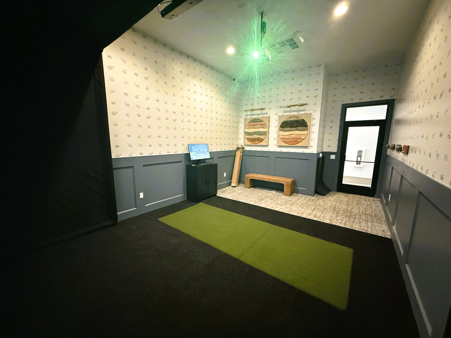 Golf simulator with green mat on floor, wall art, and computer monitor.