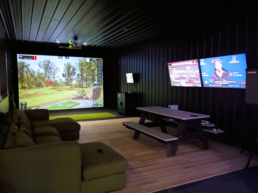 Indoor golf simulator room with two large screens displaying a golf simulation game and a sports event.