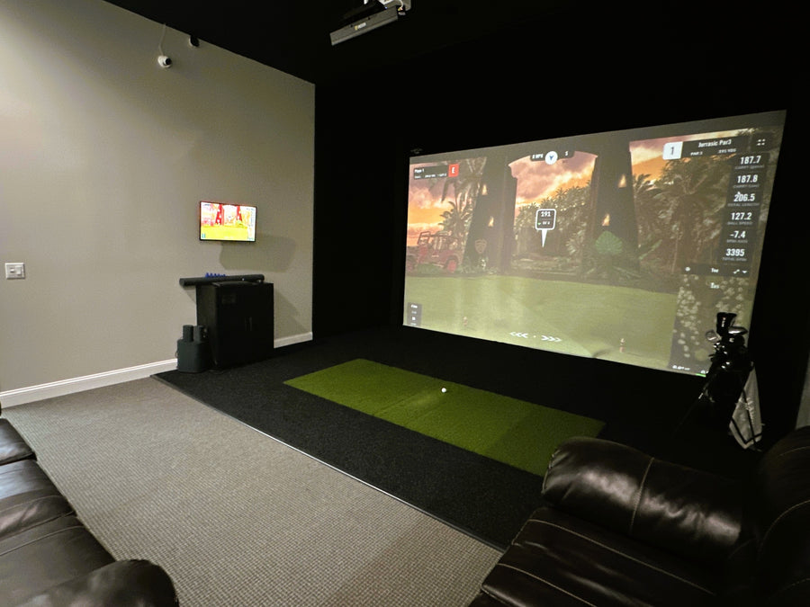 Indoor golf simulator with a large screen displaying a video game, surrounded by black leather chairs and a gray carpet.
