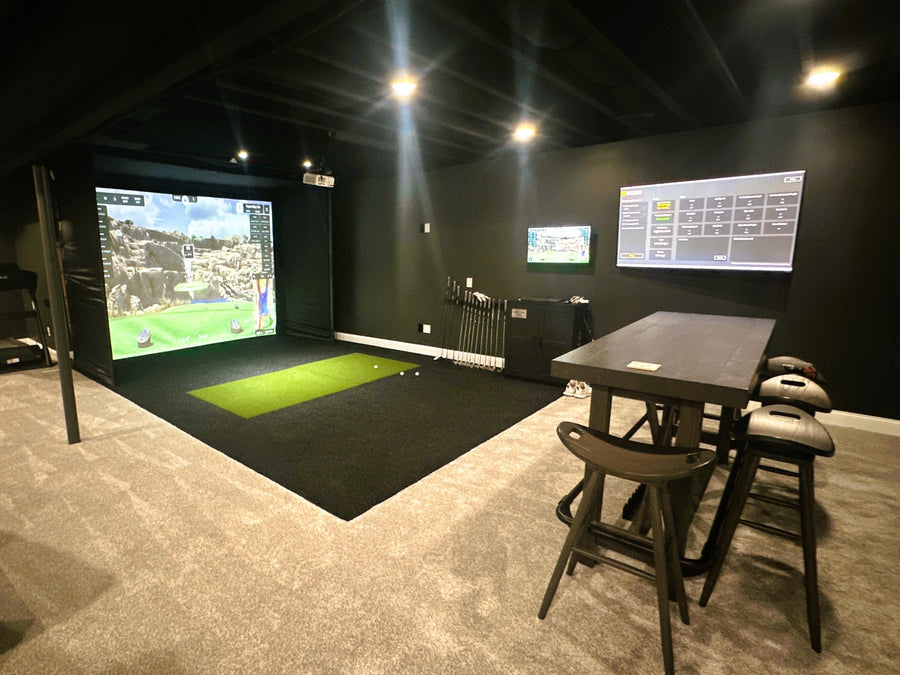 Golf simulator room with two screens, a table, and chairs in a dimly lit room.