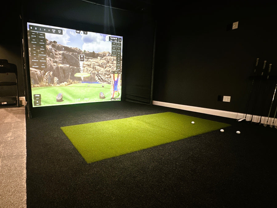 Indoor golf simulator setup with screen displaying a golf course and green mat on the floor.
