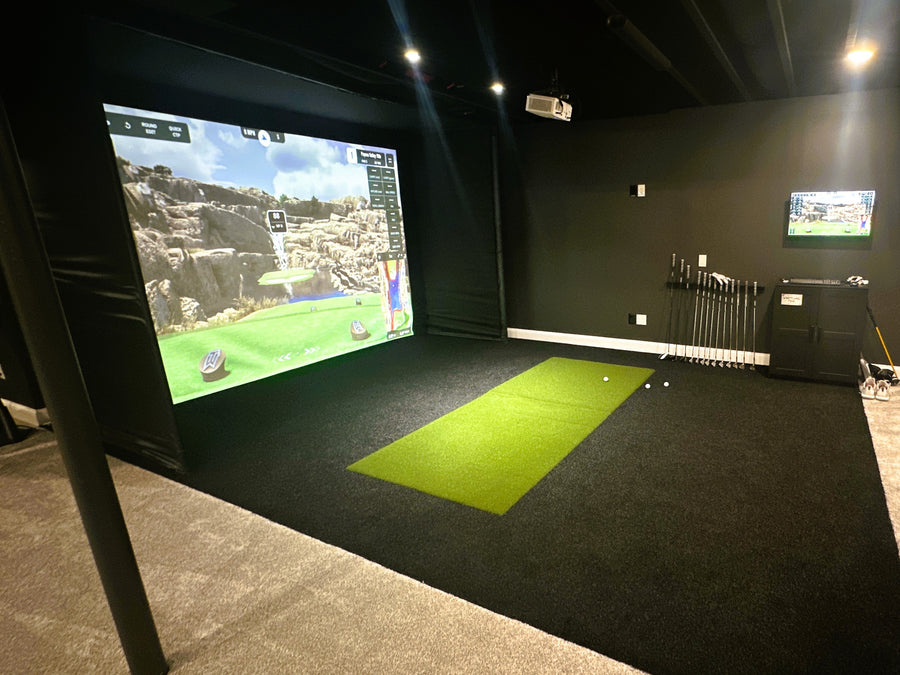 Indoor golf simulator room with green mat and screen displaying a golf course.