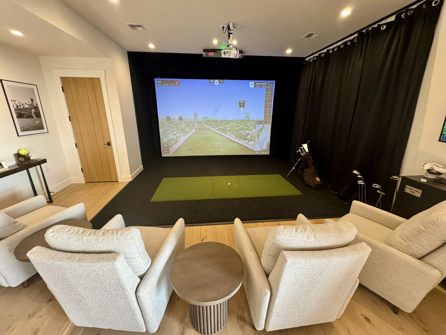 Indoor golf simulator setup in a living room with white chairs and a small table.