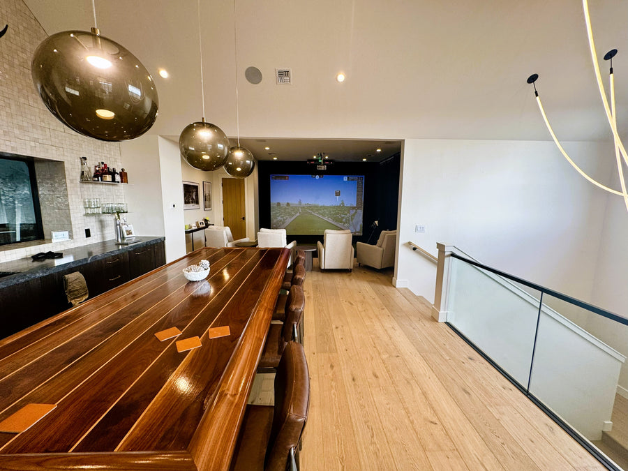 Indoor golf simulator room with a large wooden dining table and chairs, and a TV in the background.