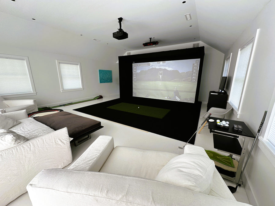 Golf simulator setup with white couches, projector, and screen displaying a nature scene.