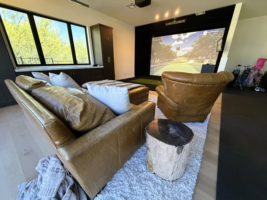 Home golf simulator setup with a large screen, brown sofa, and armchair.