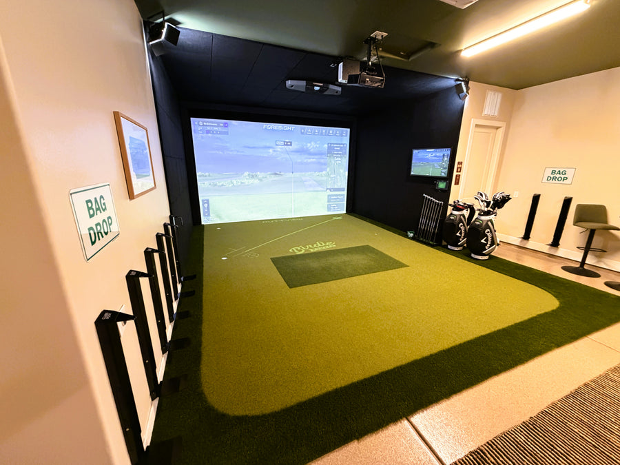 Indoor golf simulator room with green turf and large screen displaying a golf course.