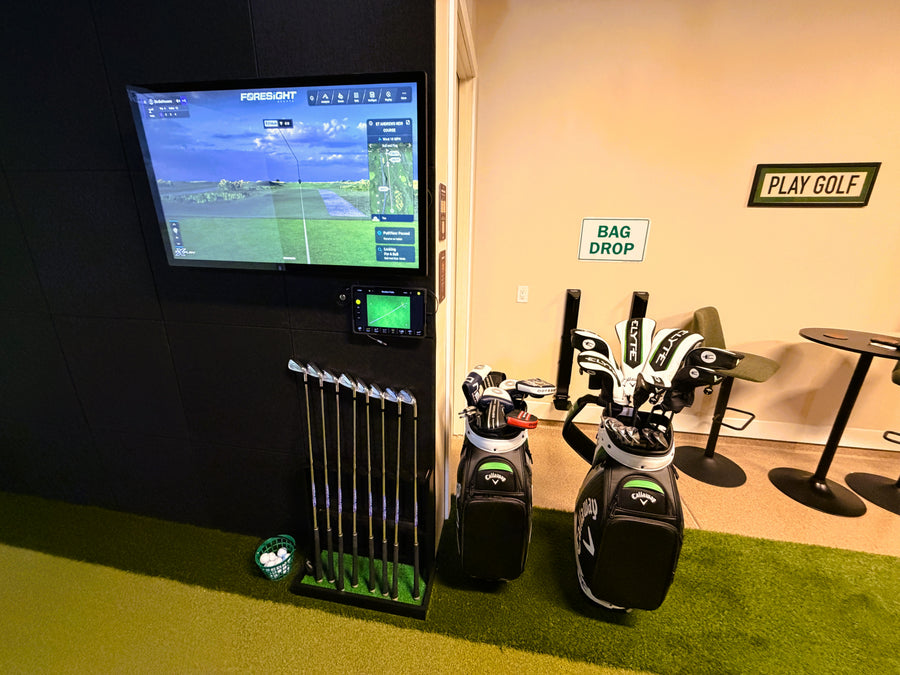 Golf simulator setup with TV screen, golf clubs, and 'Bag Drop' sign.