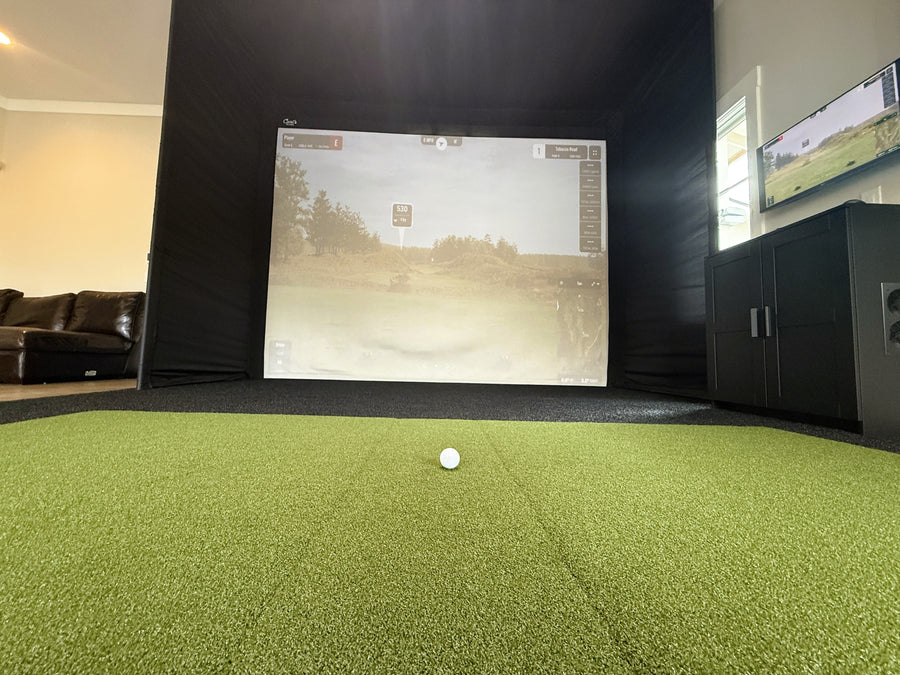 Indoor golf simulator setup with screen displaying a golf course and a ball on a green floor.