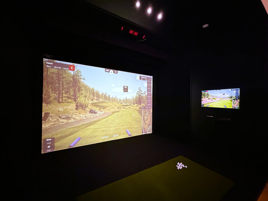 An indoor golf simulator with two screens displaying a golf game in a dark room.