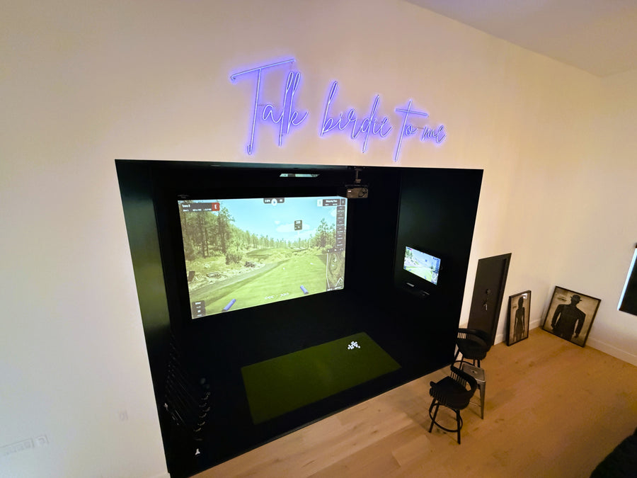Indoor golf simulator setup with two screens on a wall in a room with a neon sign.