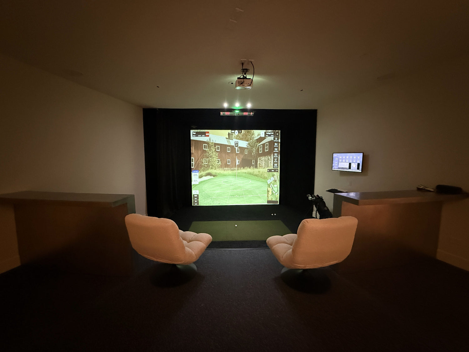 Indoor golf simulator with a screen displaying a scene, flanked by two chairs in a dimly lit room.
