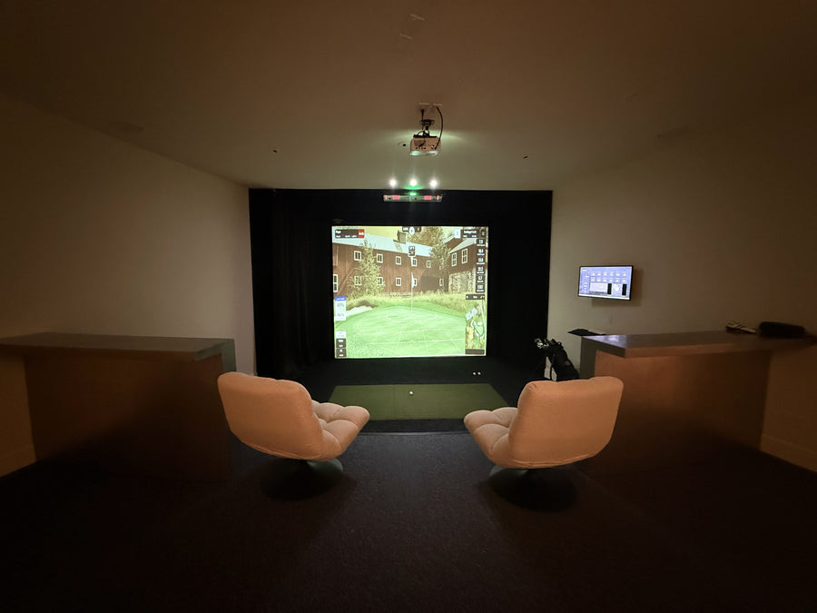 Indoor golf simulator with a screen displaying a scene, flanked by two chairs in a dimly lit room.