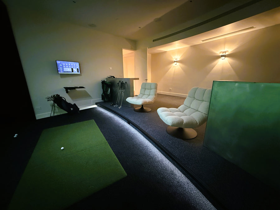 Indoor golf simulator setup with green mat, chairs, and TV in a room.