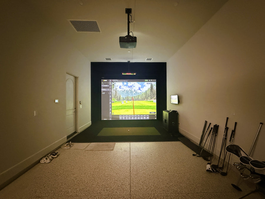 Indoor golf simulator setup with screen and golf clubs on a carpeted floor.