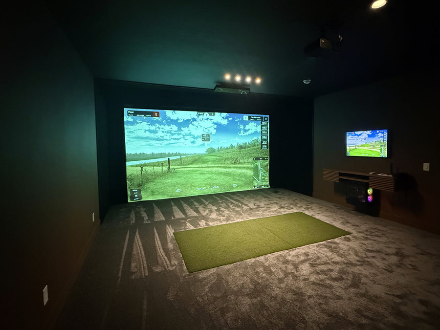 Indoor golf simulator setup with large screen and green mat on carpeted floor.
