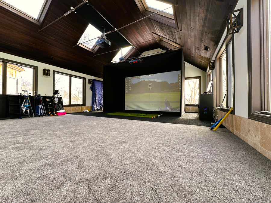 Indoor golf simulator setup with a screen displaying a golf course, surrounded by windows and skylights.