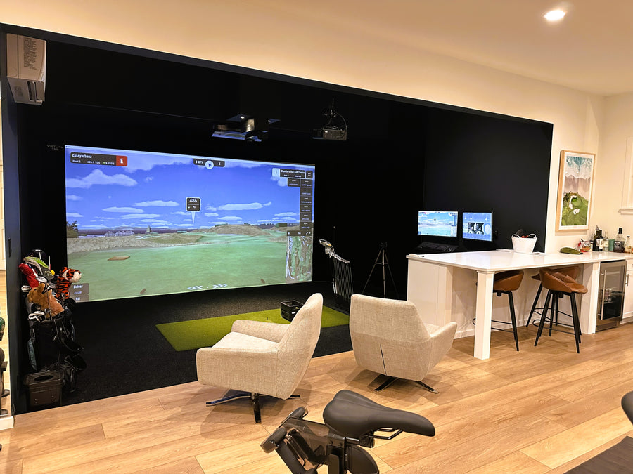 Golf Simulator room with large screen displaying a golf simulation game, surrounded by chairs and a desk.