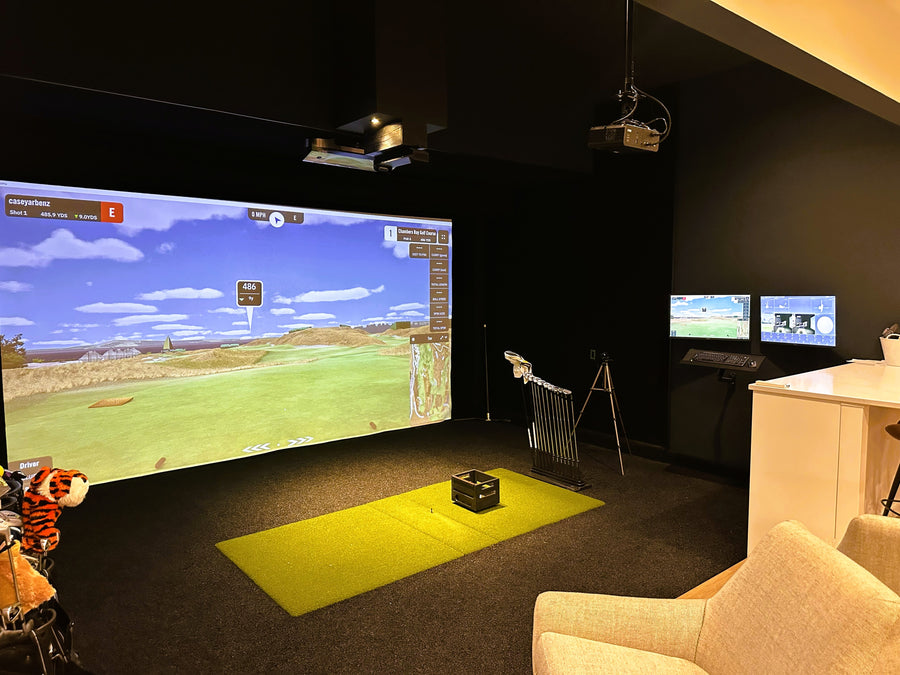 Golf simulator room with a large screen displaying a golf simulation game, surrounded by golf equipment.