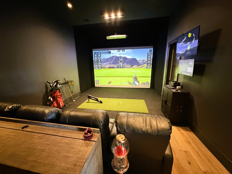 Golf simulator room with two televisions displaying a sports game, surrounded by gaming equipment.