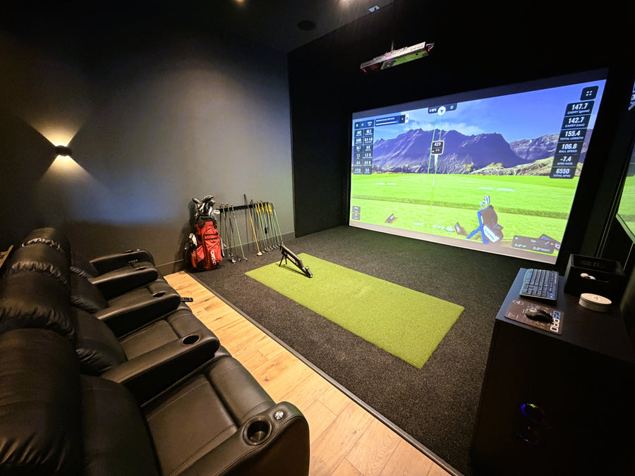 Home indoor golf simulator setup with screen displaying a golf course, surrounded by recliner chairs and golf equipment.