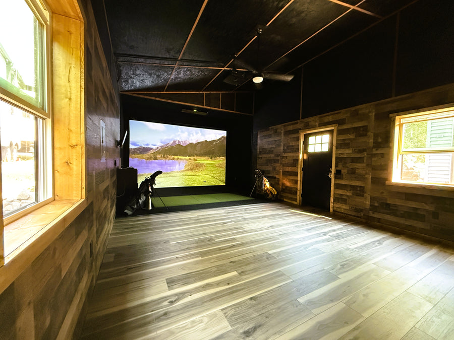 Indoor golf simulator shed with wooden walls and floor, featuring a screen displaying an outdoor scene.