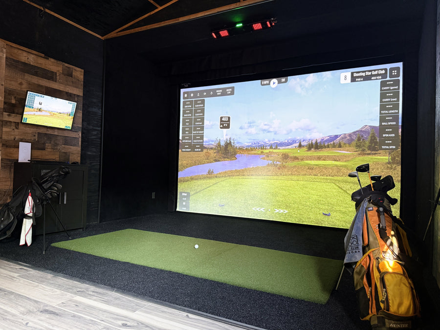Indoor golf simulator setup with screen displaying a golf course, golf clubs, and mat.