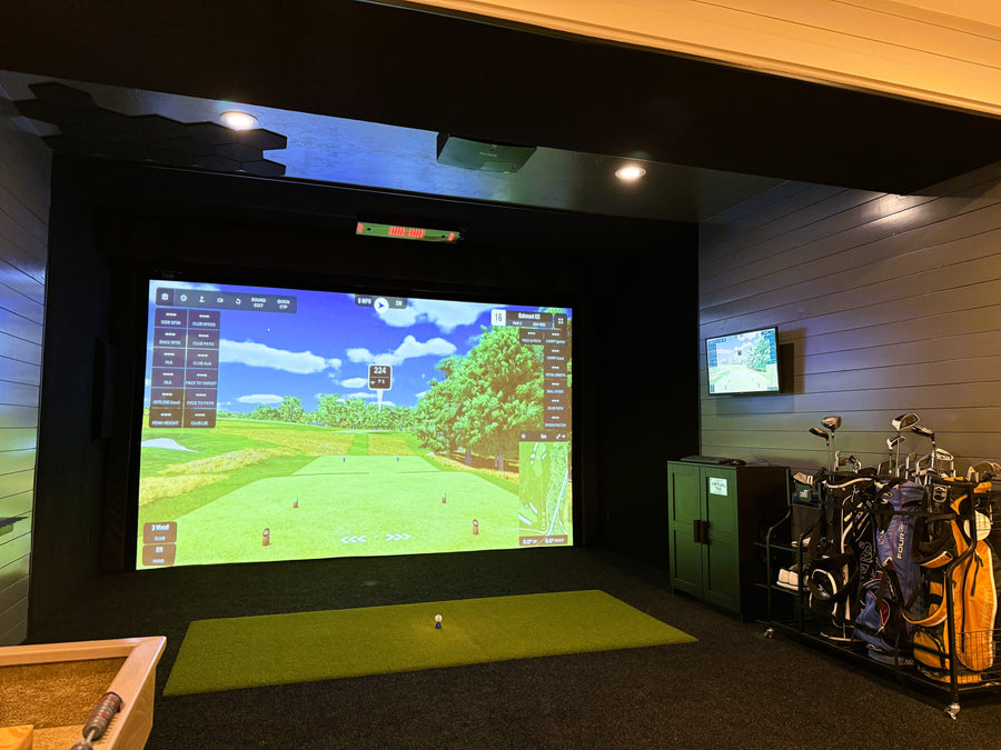 Golf simulator setup with large screen displaying a golf course, surrounded by golf bags and equipment.
