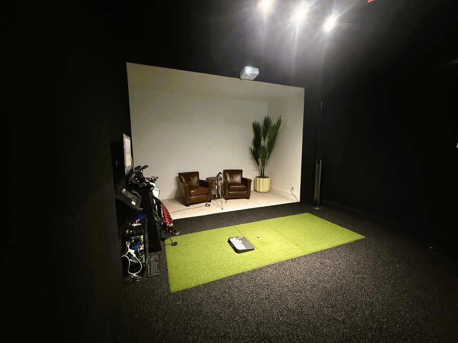 Indoor golf simulator setup with a screen displaying a room with chairs and a plant.