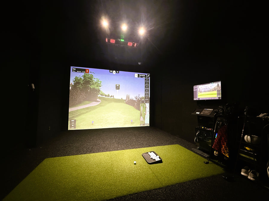 Golf simulator setup with screen displaying a golf course and various equipment on a green floor.