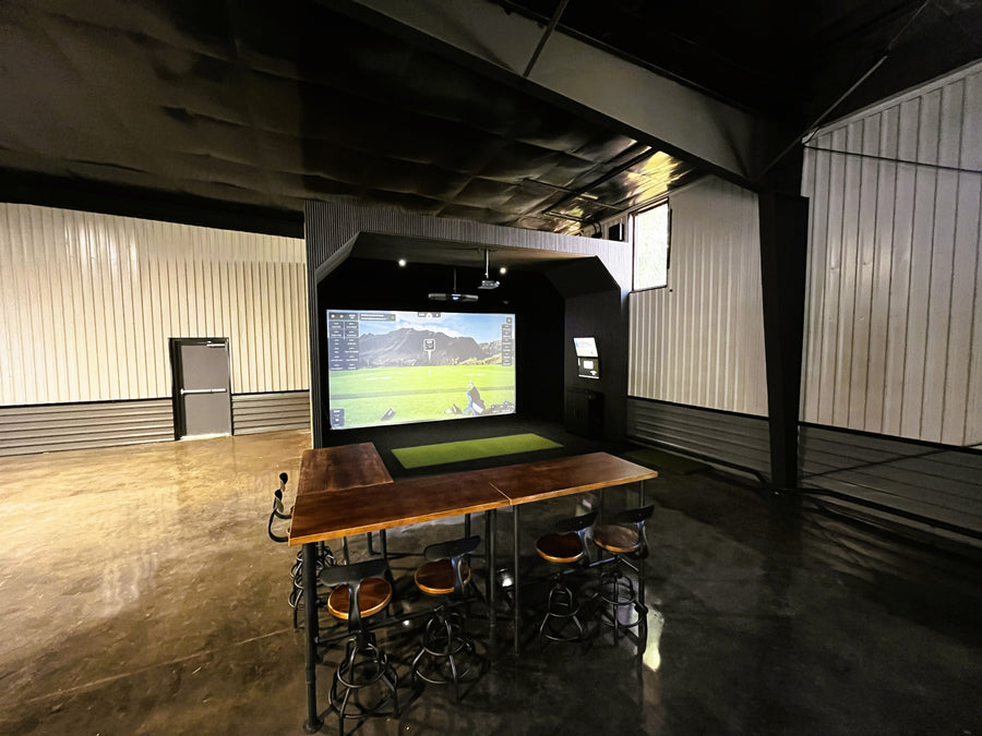 Modern indoor golf simulator with a large screen displaying a golf game, surrounded by wooden tables and chairs.