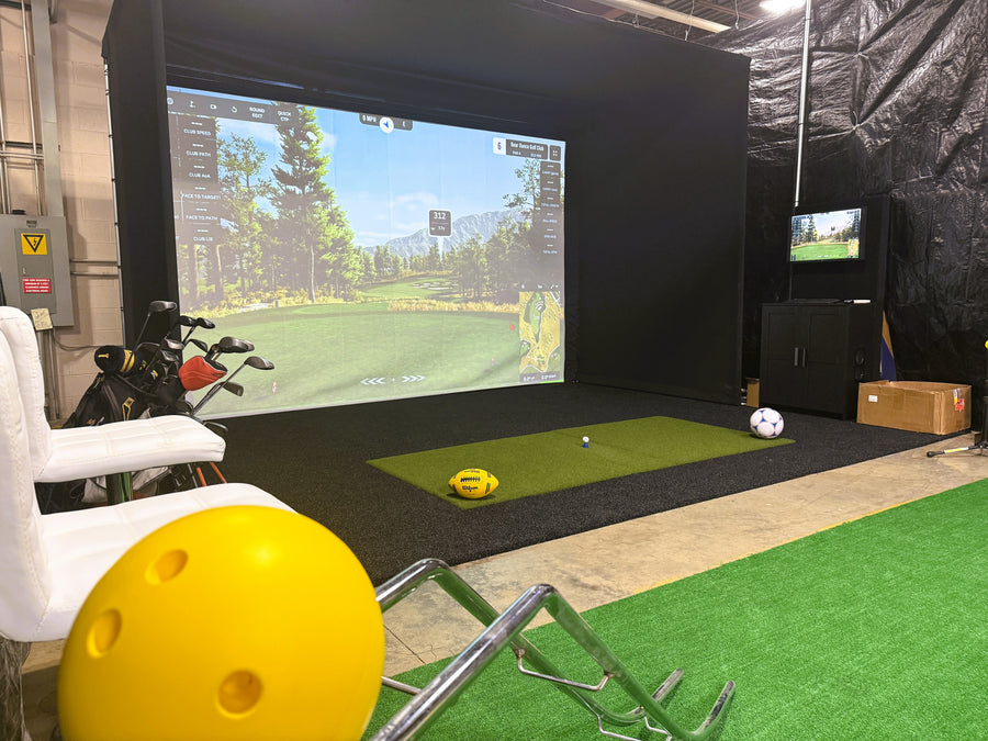 Golf simulator setup with screen displaying a golf course, golf clubs, and balls on a green floor.