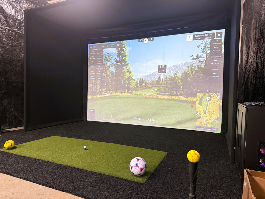 Indoor golf simulator setup with screen displaying a golf course, golf balls on a mat, and a golf club.