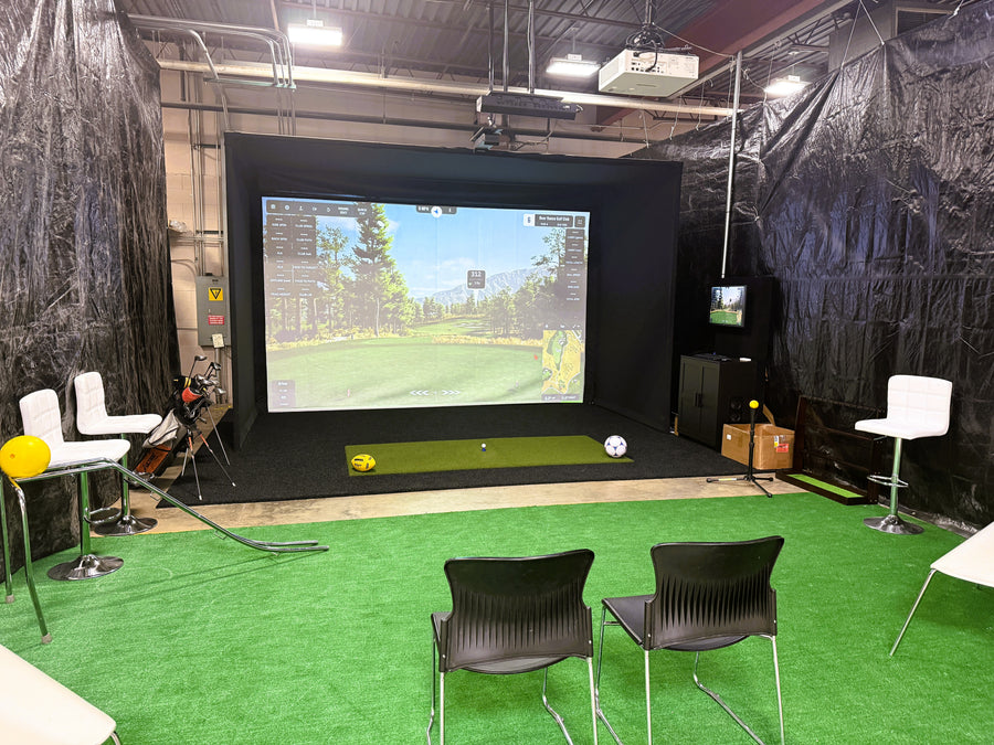 Golf simulator setup with screen, chairs, and golf equipment in a room.