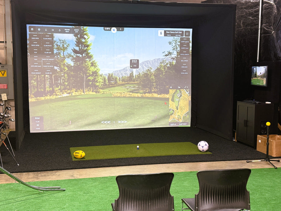 Golf simulator setup with screen displaying a golf course, two balls on a green mat, and chairs around.