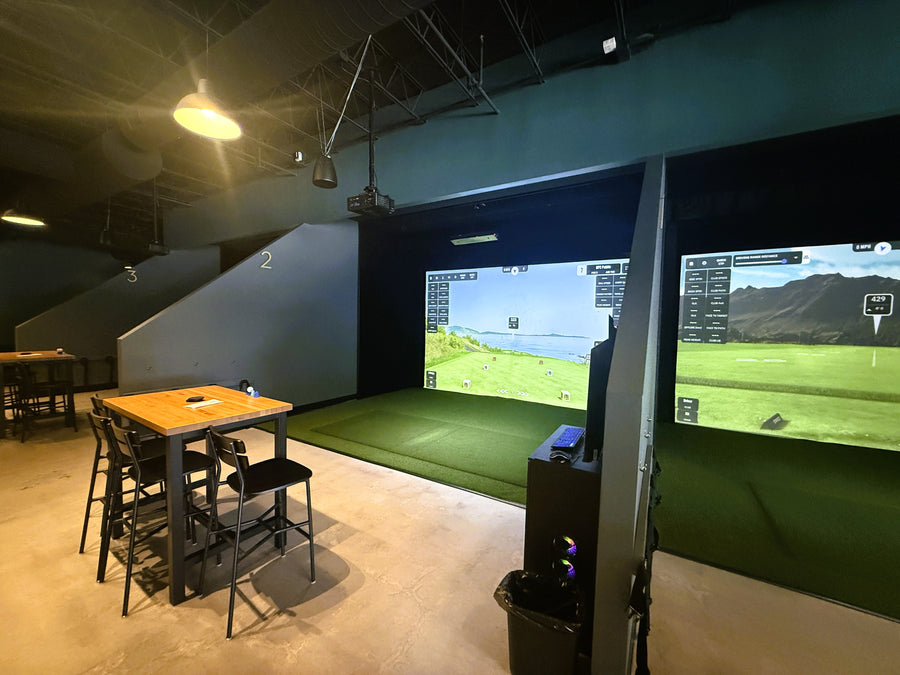 Golf simulator setup with screen displaying a golf course, surrounded by chairs and tables.