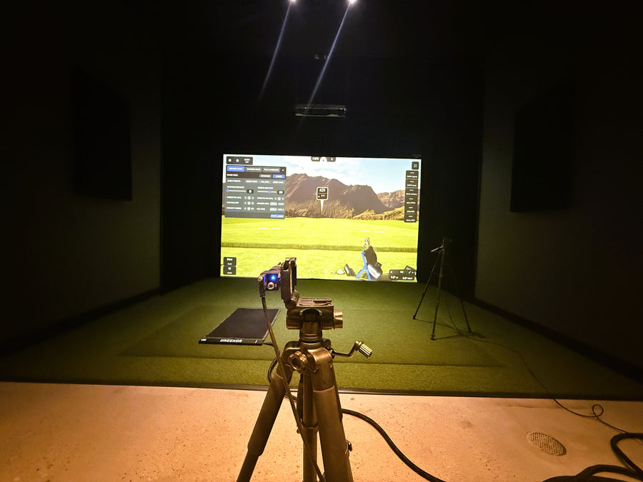Golf simulator setup with screen displaying a golf course and equipment on a grassy area.