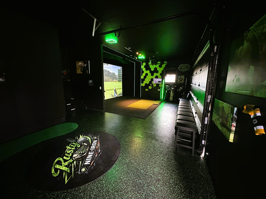 Indoor golf simulator room with multiple screens and a green logo on the floor.