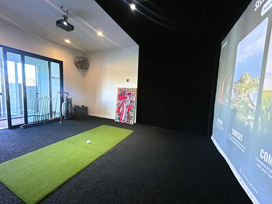 Indoor golf simulator setup with a screen displaying a golf course, a green mat on the floor, and various equipment.