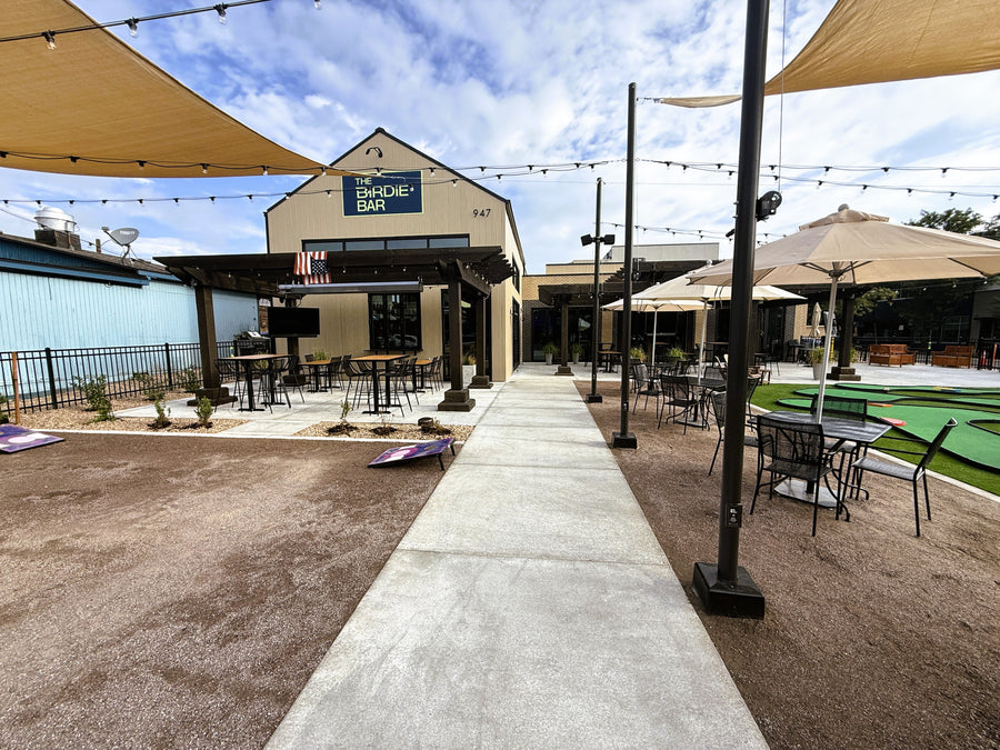 Outdoor patio area with tables, chairs, and umbrellas in front of a building with 'The Birdie Bar' sign.