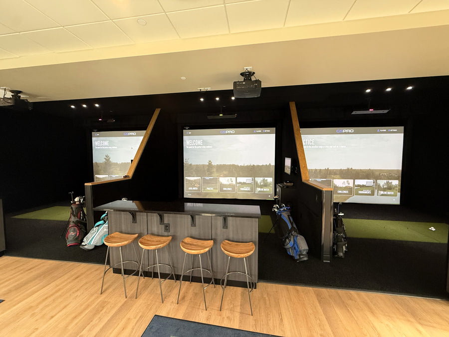Golf simulator room with multiple screens and stools.