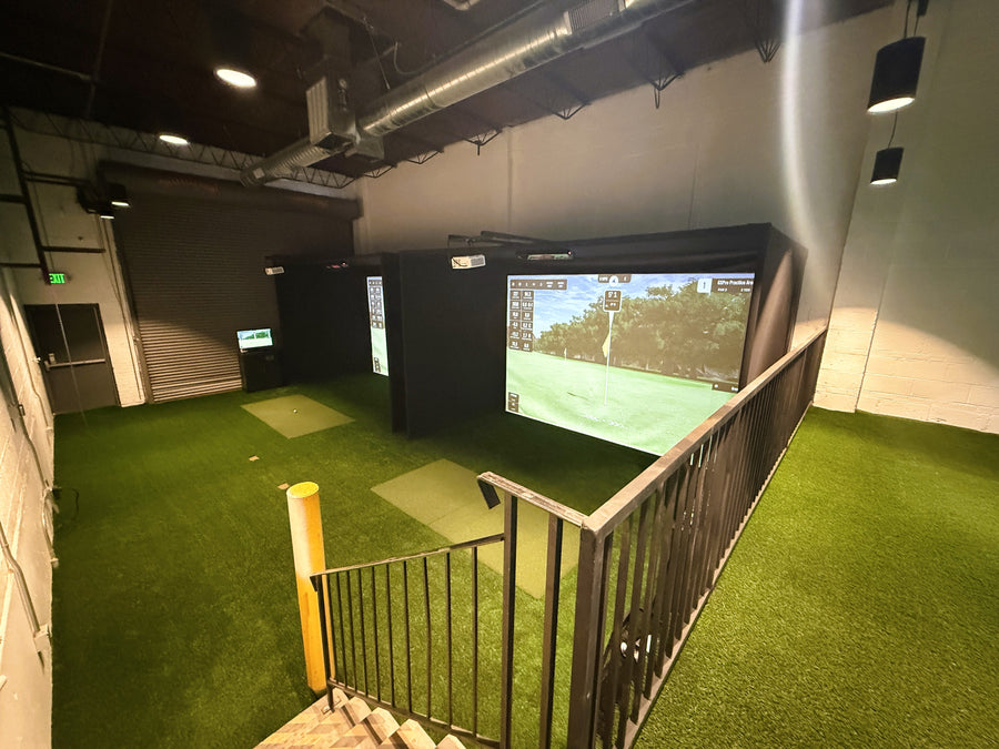 Indoor golf simulator setup with green turf and screen displaying a golf course.