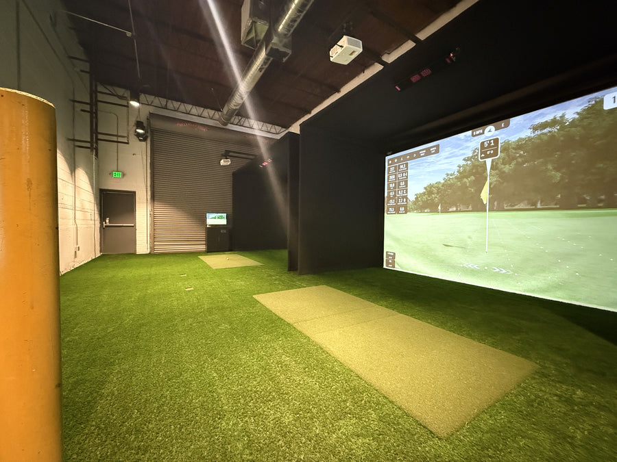 Indoor golf simulator with green turf and large screen displaying a golf course.