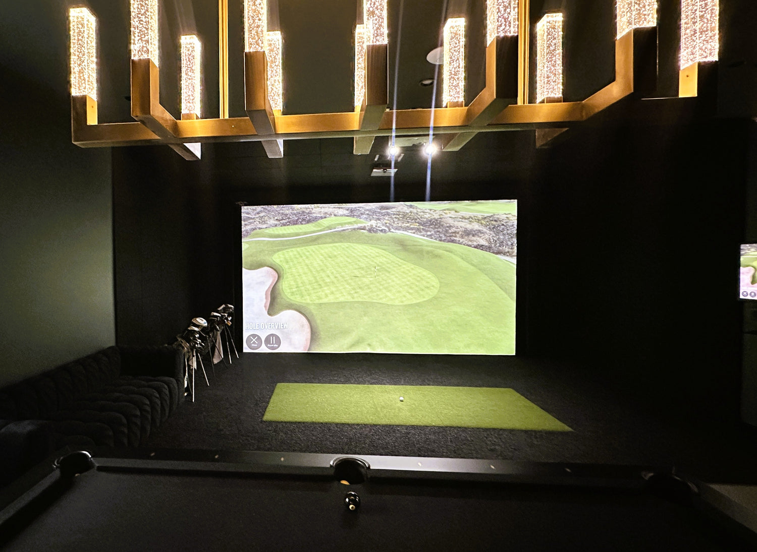 Indoor golf simulator setup with screen displaying a golf course, golf clubs, and mat.