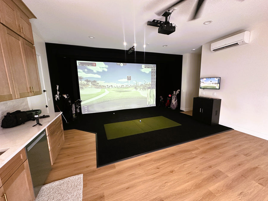 Golf simulator with a large screen displaying a sports game, surrounded by wooden cabinets and a kitchen area.