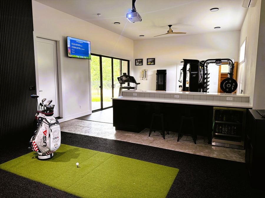 Indoor golf simulator setup with green mat, golf bags, and TV screen.