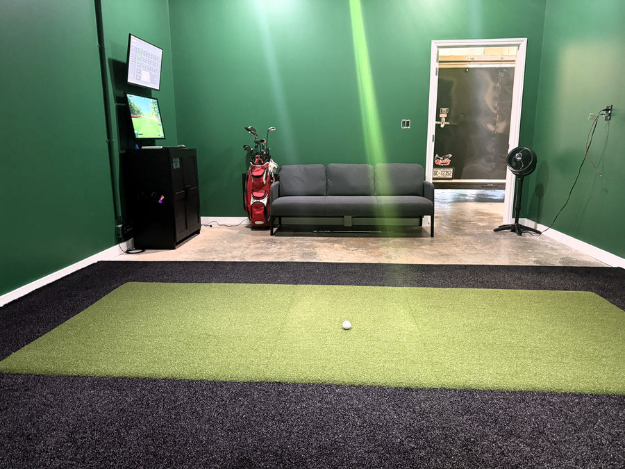 Indoor golf practice area with green mat, ball, and equipment against a green wall.
