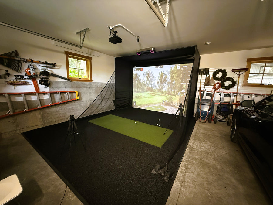 Garage golf simulator with tools and ladder on the wall. Screen displaying round of golf with green turf on the ground.