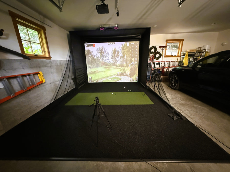 Garage golf simulator with tools and ladder on the wall. Screen displaying round of golf with green turf on the ground.
