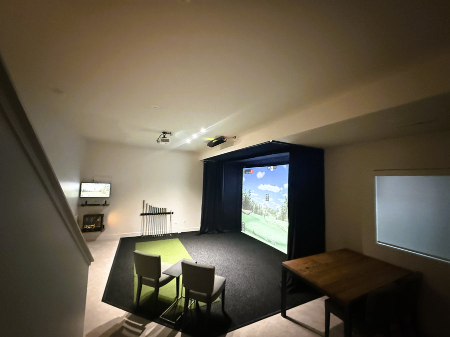 Home golf simulator setup with a screen displaying a sports game, surrounded by chairs and a small table.